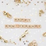 The words 'HAPPY ANNIVERSARY' in wooden letter tiles, surrounded by gold and silver coloured stars and gold curls, all set against a white background.