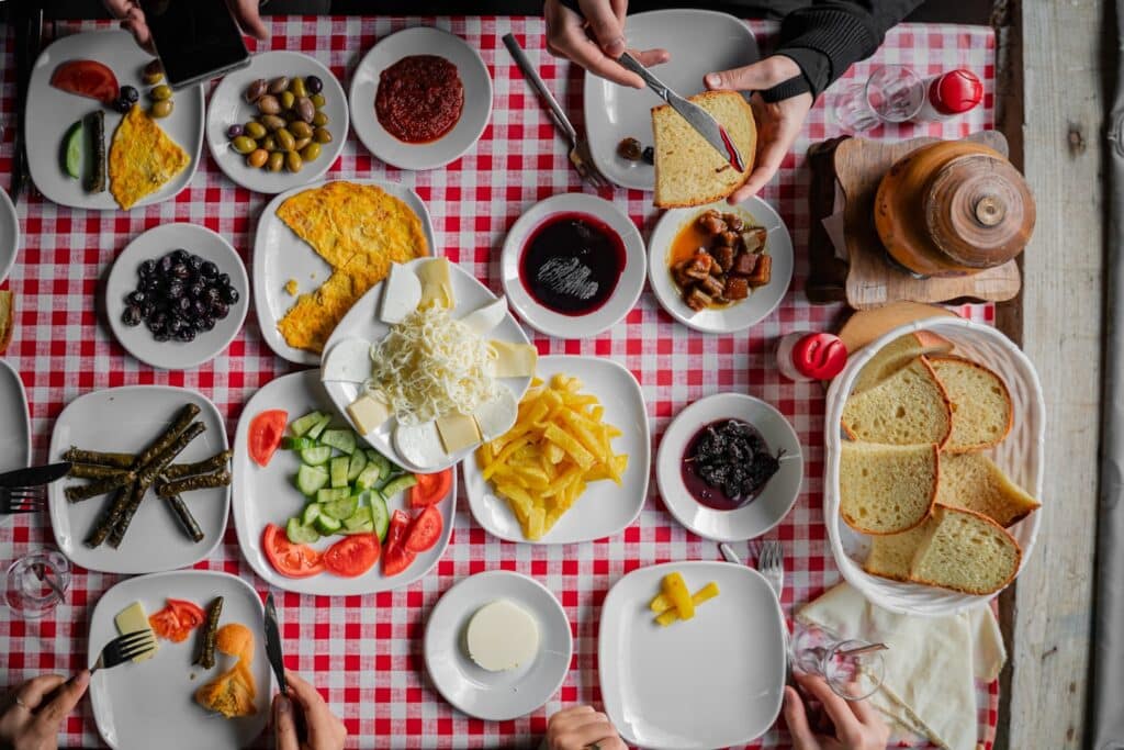 An overhead view of a traditional Turkish breakfast table filled with cheese, olives, bread, eggs, jams, and tea, shared by multiple people around a red checkered tablecloth.