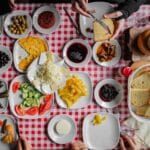 An overhead view of a traditional Turkish breakfast table filled with cheese, olives, bread, eggs, jams, and tea, shared by multiple people around a red checkered tablecloth.