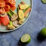 An overhead shot of A vibrant top-down shot of shrimp in a red sauce next to perfectly blanched vegetables (baby corn, mushrooms) on a light blue plate, garnished with lime and basil.